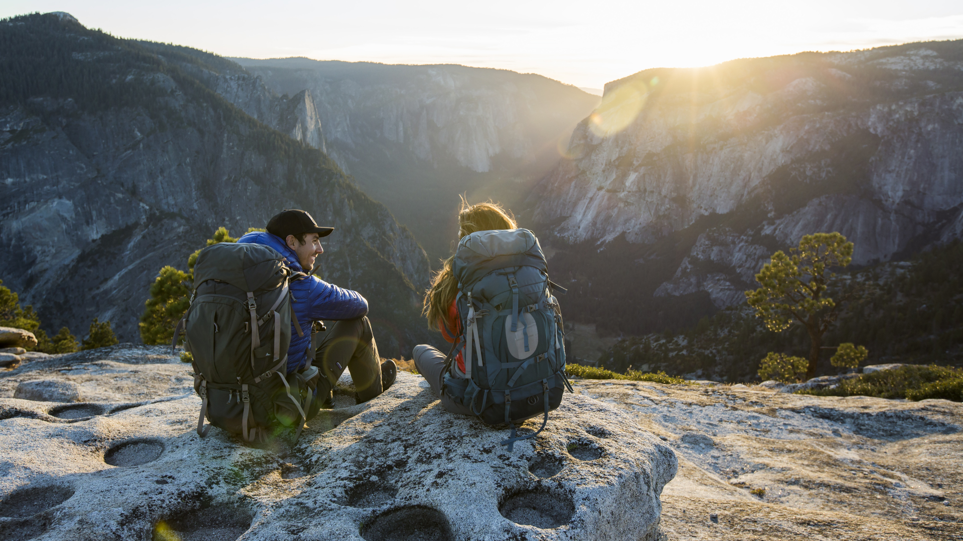 Un hombre y una mujer se sientan en un acantilado en Yosemite con mochilas y mirando la vista