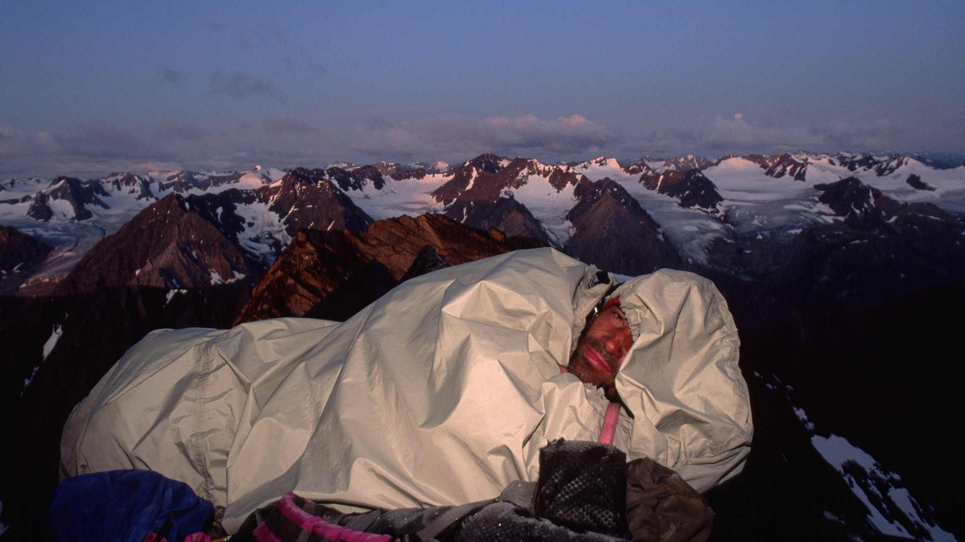 Un hombre durmiendo en un saco bivy con montañas nevadas al fondo