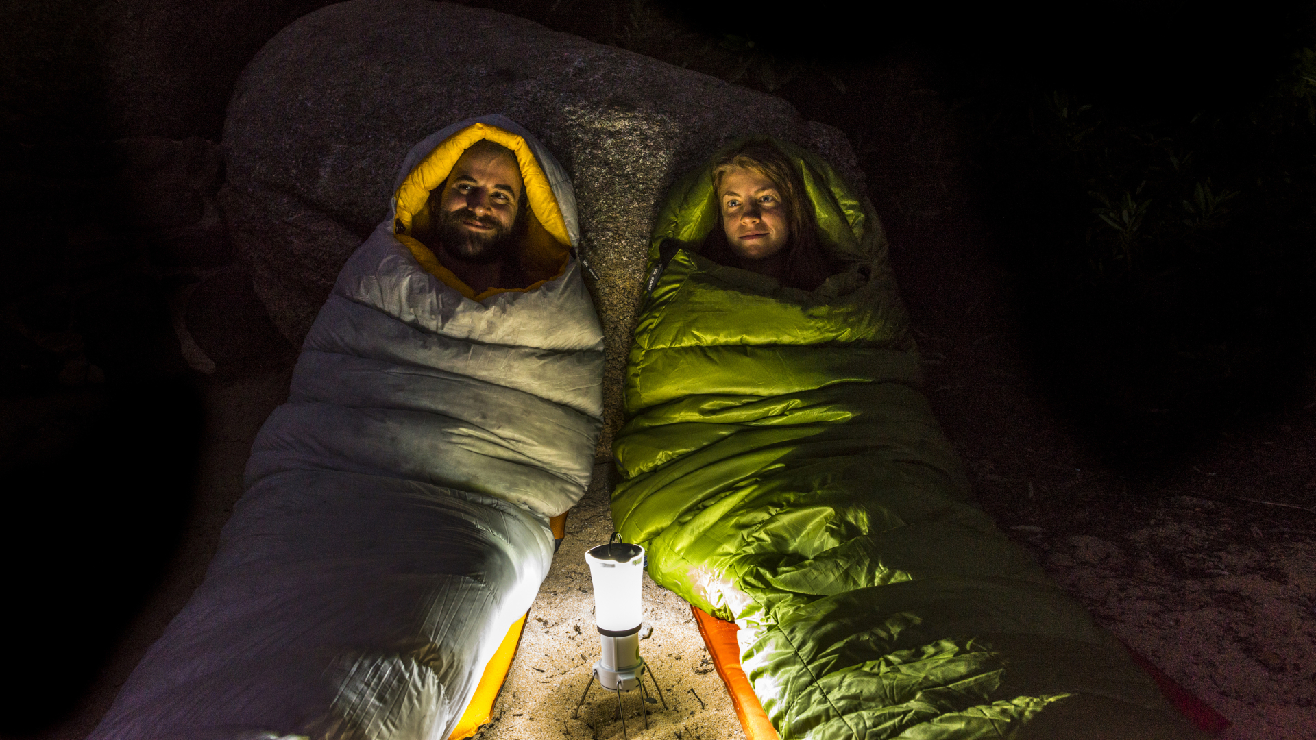 Hombre y mujer en sacos de dormir sonriendo iluminados por una linterna por la noche.